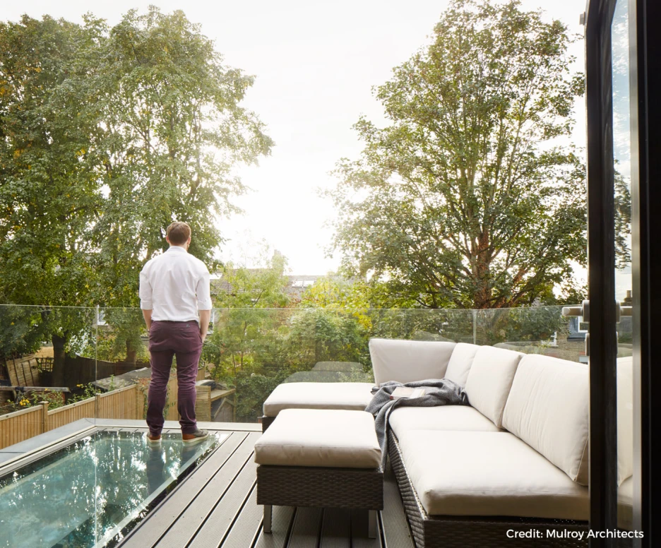 A person stands on a balcony overlooking a lush green landscape. The balcony features a glass railing and a cozy seating area with light-colored cushions. The sky is bright, indicating daylight.