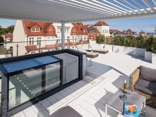 A modern rooftop terrace featuring a wooden deck, lounge furniture, and a view of nearby buildings with red rooftops under a clear blue sky. A table with a bottle of wine and a colorful artwork is displayed.