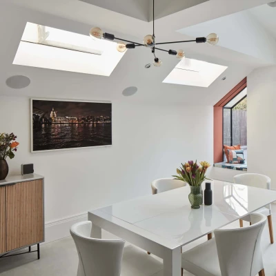 Bright modern dining area featuring a white table surrounded by four chairs. Skylights illuminate the space, complemented by a wall painting and a sideboard with a floral arrangement. A glass door leads to an outdoor patio.