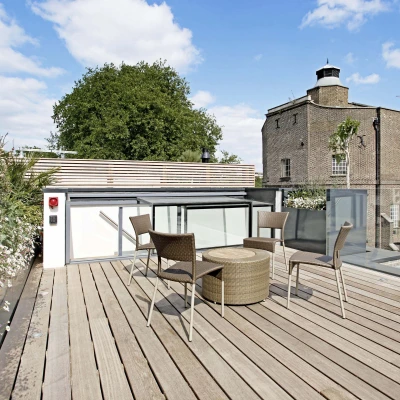 A modern rooftop terrace features a wooden deck with a round table and four chairs. Lush flowers border the area, and a brick building stands in the background under a partly cloudy sky.