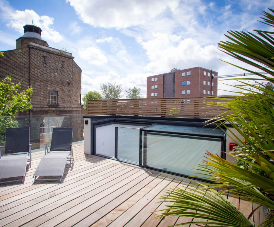 A modern rooftop terrace featuring two lounge chairs, surrounded by greenery. In the background, a historic brick building and a taller apartment building are visible under a partly cloudy sky.