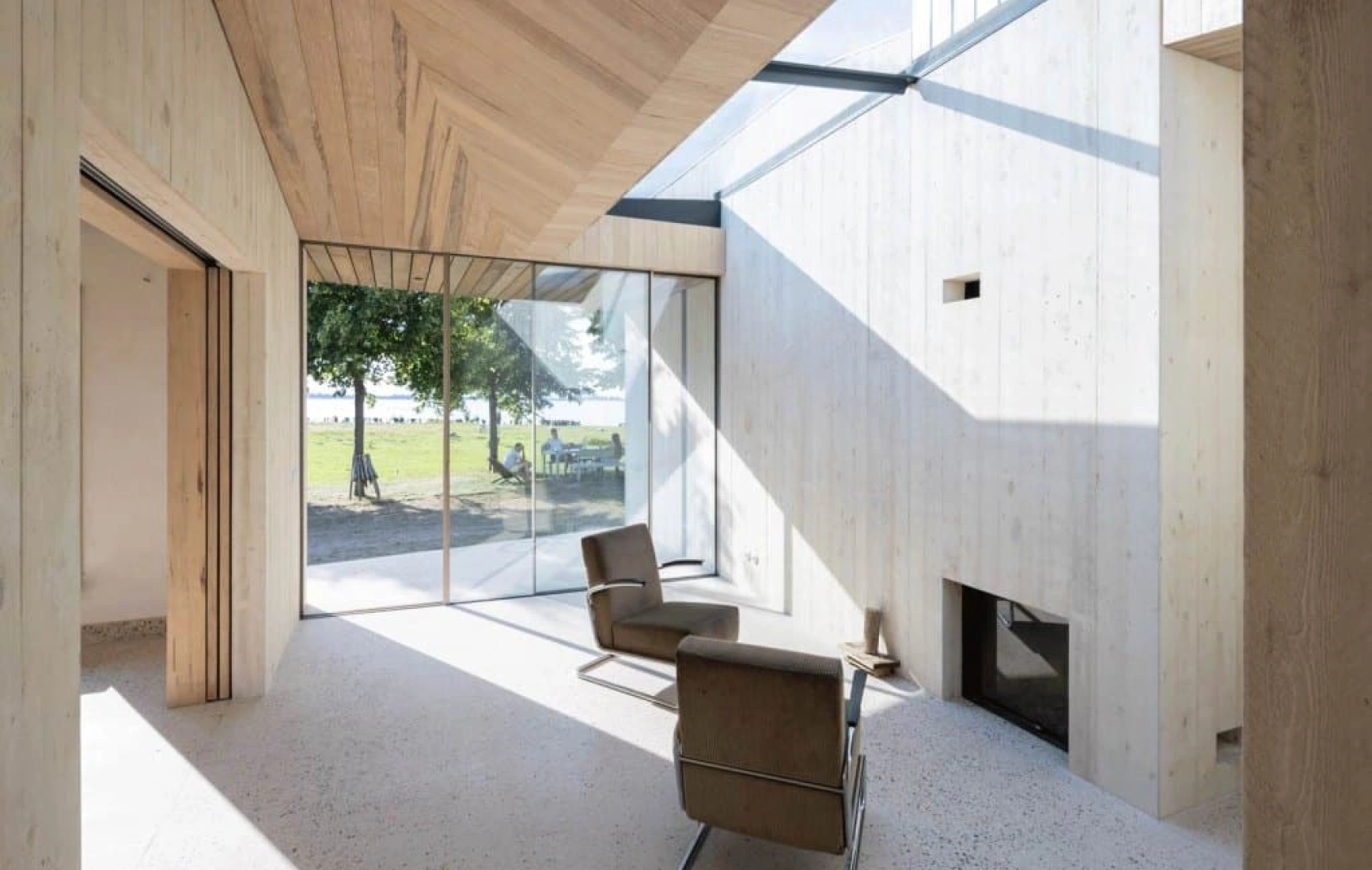 A modern interior space featuring wooden walls and ceiling, large glass windows allowing natural light in, and a minimalist chair. Visible outside are trees and a grassy area.