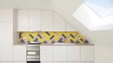 Modern kitchen featuring sleek white cabinetry, a stainless steel stove, and a colorful herringbone-patterned backsplash in shades of yellow, pink, and gray. A skylight adds natural light to the space.