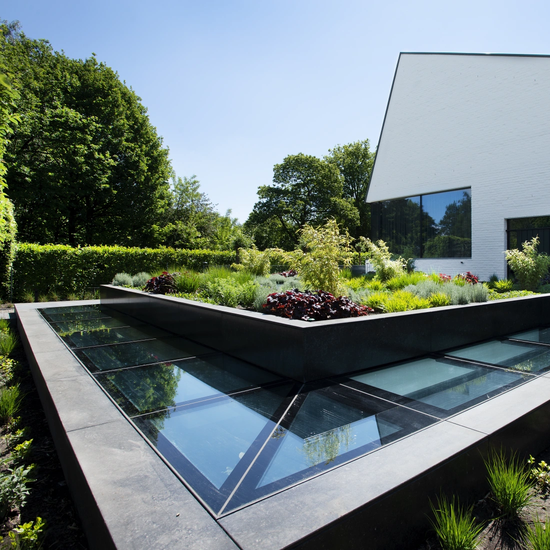 A modern building with a unique angular design is surrounded by a landscaped garden featuring lush greenery and colorful plants. The garden includes a geometric raised bed with glass panels.
