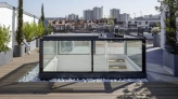 A modern rooftop space featuring a glass enclosure with wooden steps, surrounded by planters and white pebbles. In the background, urban buildings and a clear blue sky are visible.