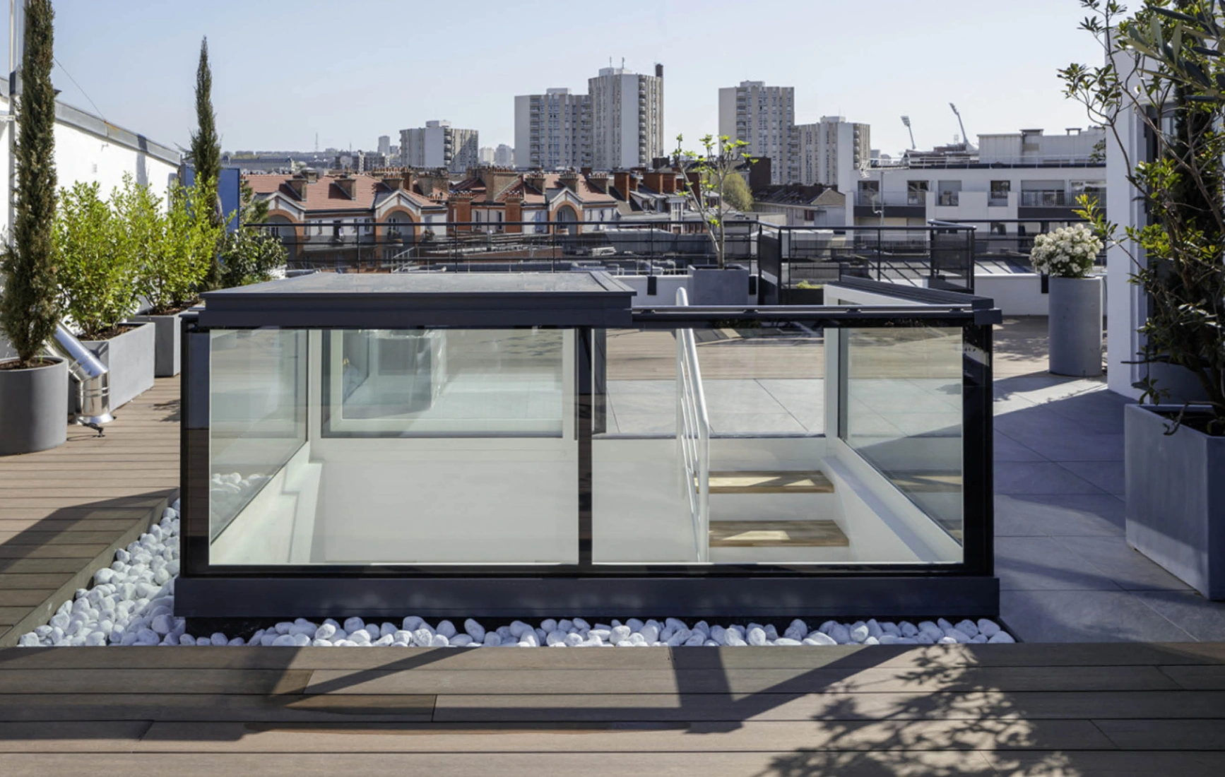 A modern rooftop space featuring a glass enclosure with wooden steps, surrounded by planters and white pebbles. In the background, urban buildings and a clear blue sky are visible.