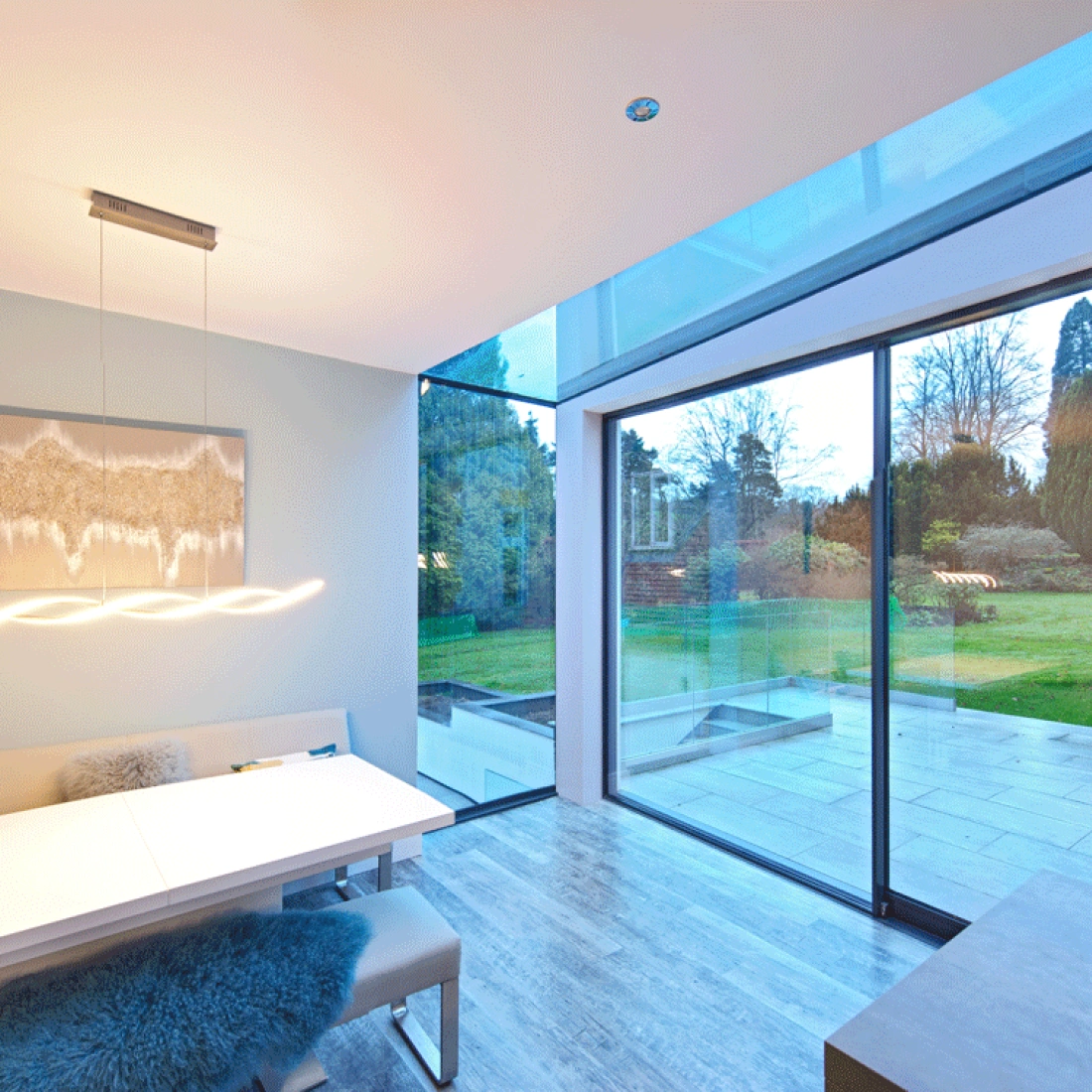 A modern dining area featuring a sleek white table and soft seating, with a large glass wall providing a view of a lush garden outside. Soft lighting hangs above the table, creating a warm atmosphere.