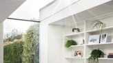 Bright, modern interior featuring a cozy chair by a window, with built-in white shelving adorned with plants and decorative items. Sunlight streams through a skylight, illuminating the space. A lush garden is visible outside.