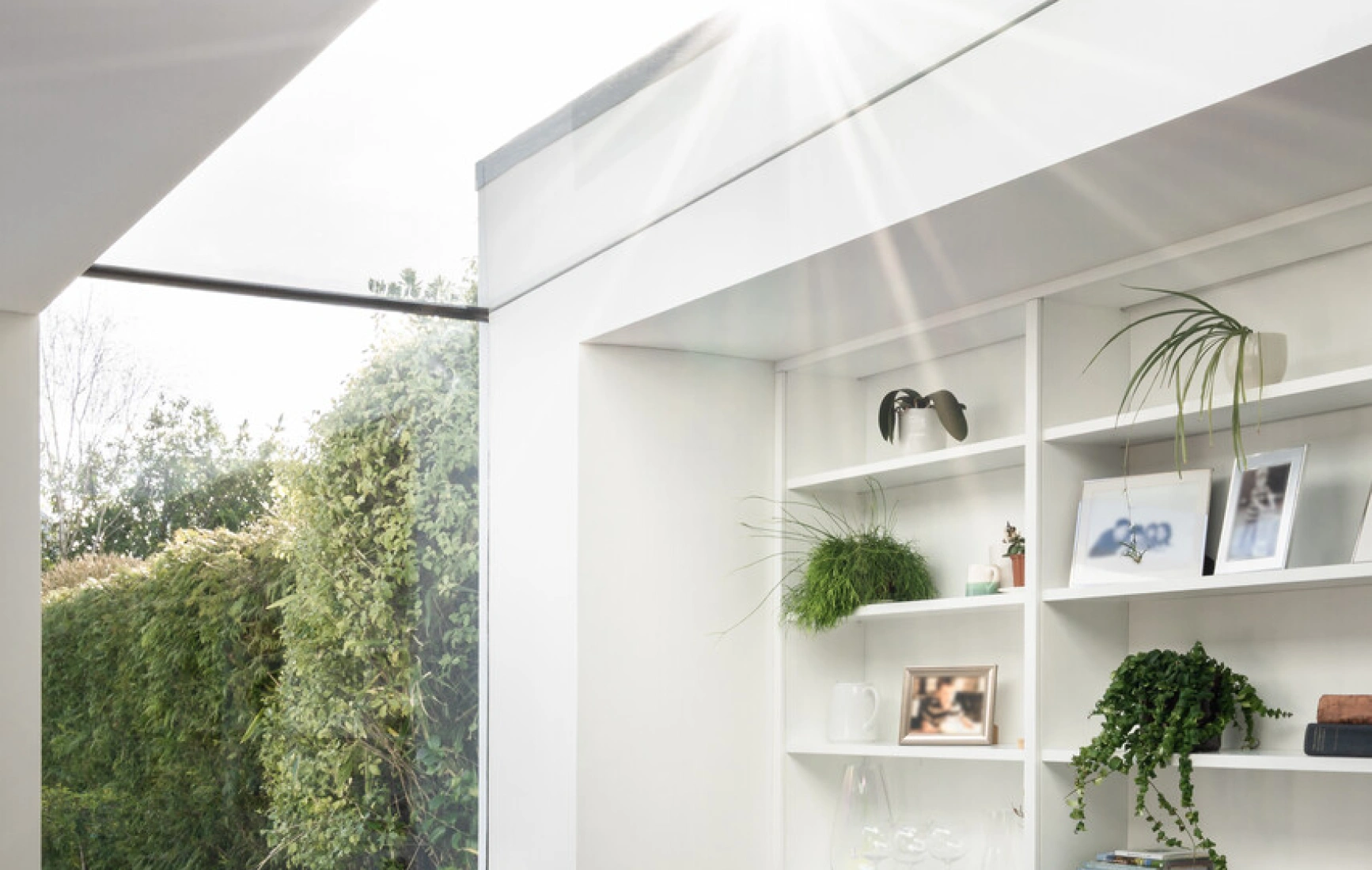 Bright, modern interior featuring a cozy chair by a window, with built-in white shelving adorned with plants and decorative items. Sunlight streams through a skylight, illuminating the space. A lush garden is visible outside.