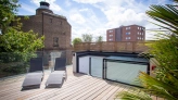 A rooftop terrace features two lounge chairs on wooden decking, surrounded by greenery. In the background, a historic brick building and a modern apartment complex are visible under a partly cloudy sky.