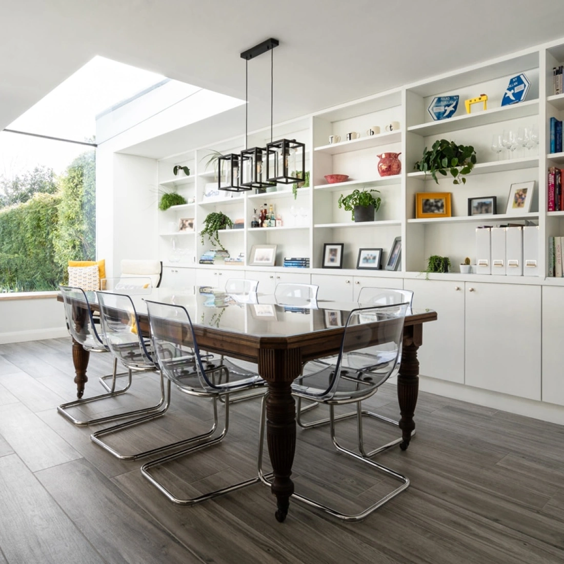 A modern kitchen and dining area features a long wooden table surrounded by transparent chairs. A large skylight above illuminates the space, highlighting built-in shelves filled with plants and colorful books. A cozy window seat is visible in the background.