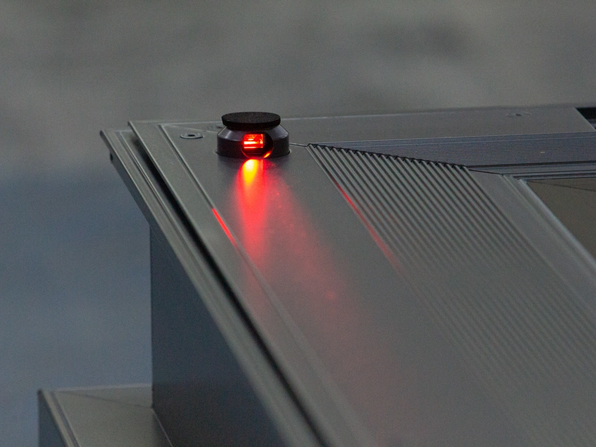 A close-up view of a device with a small, round black button featuring a glowing red light. The button is positioned on a metallic surface with ridges and a sleek design.