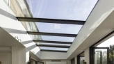 Bright interior space with a glass ceiling and large windows. Sunlight casts shadows on the wooden floor, creating a warm atmosphere. Modern design includes open doors leading to an outdoor area.