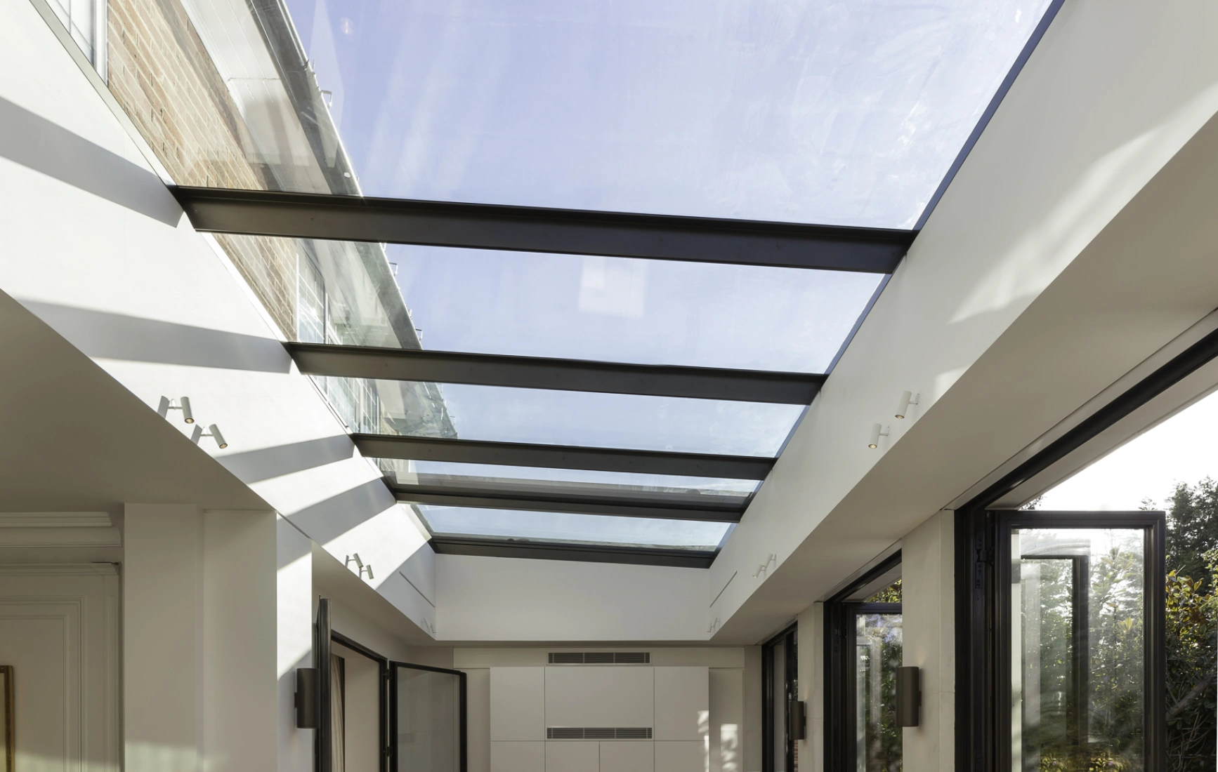 Bright interior space with a glass ceiling and large windows. Sunlight casts shadows on the wooden floor, creating a warm atmosphere. Modern design includes open doors leading to an outdoor area.