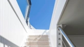 A well-lit staircase with soft beige carpet leading upward, flanked by sleek white walls and a modern railing. The bright blue sky is visible through large windows at the top.