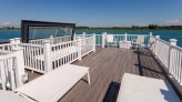 A spacious wooden deck with white lounge chairs and railings overlooks a calm blue body of water, under a clear blue sky.