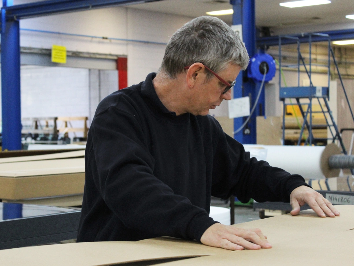 A man with gray hair wearing glasses is working at a table in a packaging facility, focusing on folding a large cardboard sheet. The background shows industrial equipment and workspace setups.