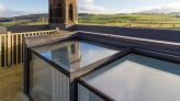 A view from a rooftop terrace featuring a modern glass structure reflecting the sky, with a tall, pointed church spire in the background against a blue sky and rolling green hills.