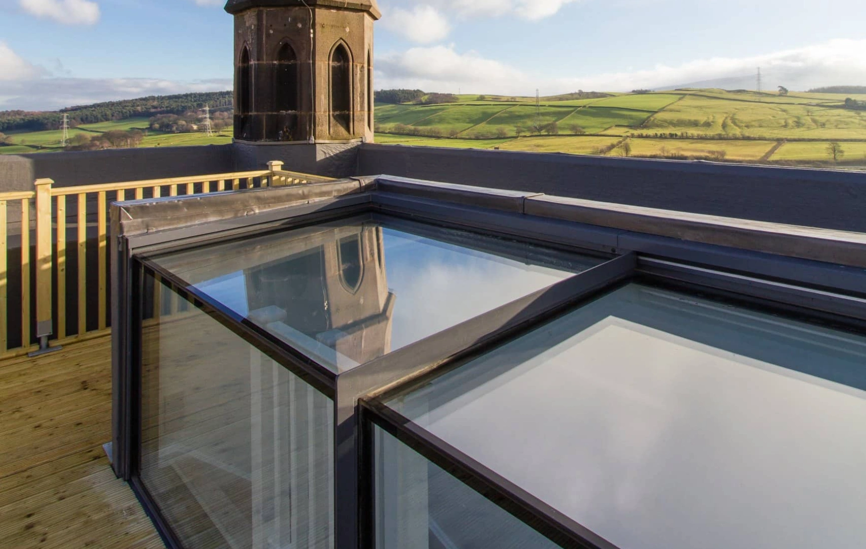 A view from a rooftop terrace featuring a modern glass structure reflecting the sky, with a tall, pointed church spire in the background against a blue sky and rolling green hills.