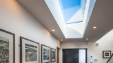 A modern hallway featuring a dark front door, framed artwork on the walls, and a wooden console table with a white lamp and yellow flowers. Natural light streams in through a skylight above.