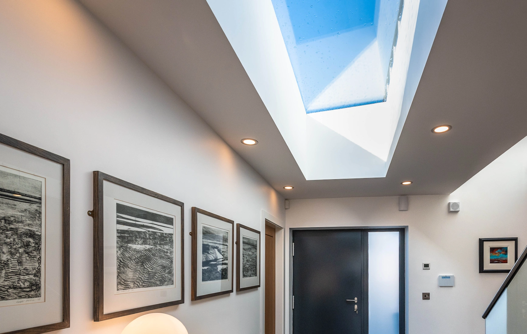 A modern hallway featuring a dark front door, framed artwork on the walls, and a wooden console table with a white lamp and yellow flowers. Natural light streams in through a skylight above.