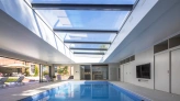 A modern indoor swimming pool with a glass ceiling, featuring sleek white walls and a flat-screen TV mounted on the wall. Sunlight streams through the skylights, illuminating the water. Comfortable lounge chairs are arranged around the pool area.