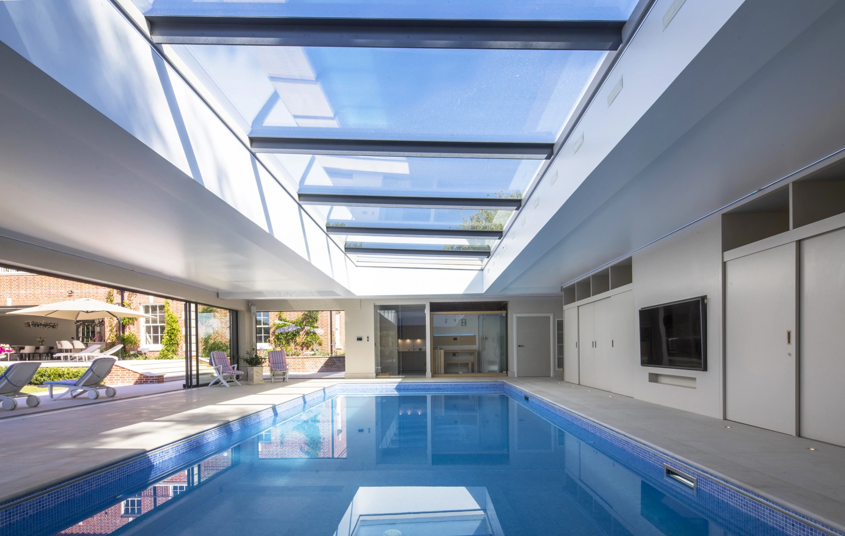A modern indoor swimming pool with a glass ceiling, featuring sleek white walls and a flat-screen TV mounted on the wall. Sunlight streams through the skylights, illuminating the water. Comfortable lounge chairs are arranged around the pool area.