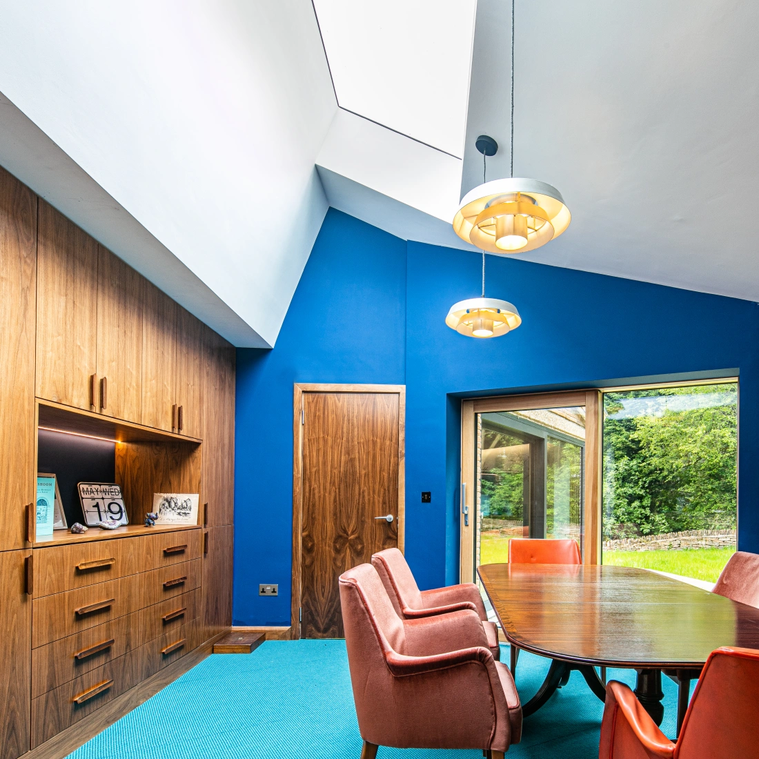 A modern dining area featuring a round wooden table surrounded by six pink upholstered chairs. The walls are painted a vibrant blue, accented by wooden cabinetry. A large window offers a view of greenery outside, and a sunburst wall clock and pendant lights add a stylish touch.
