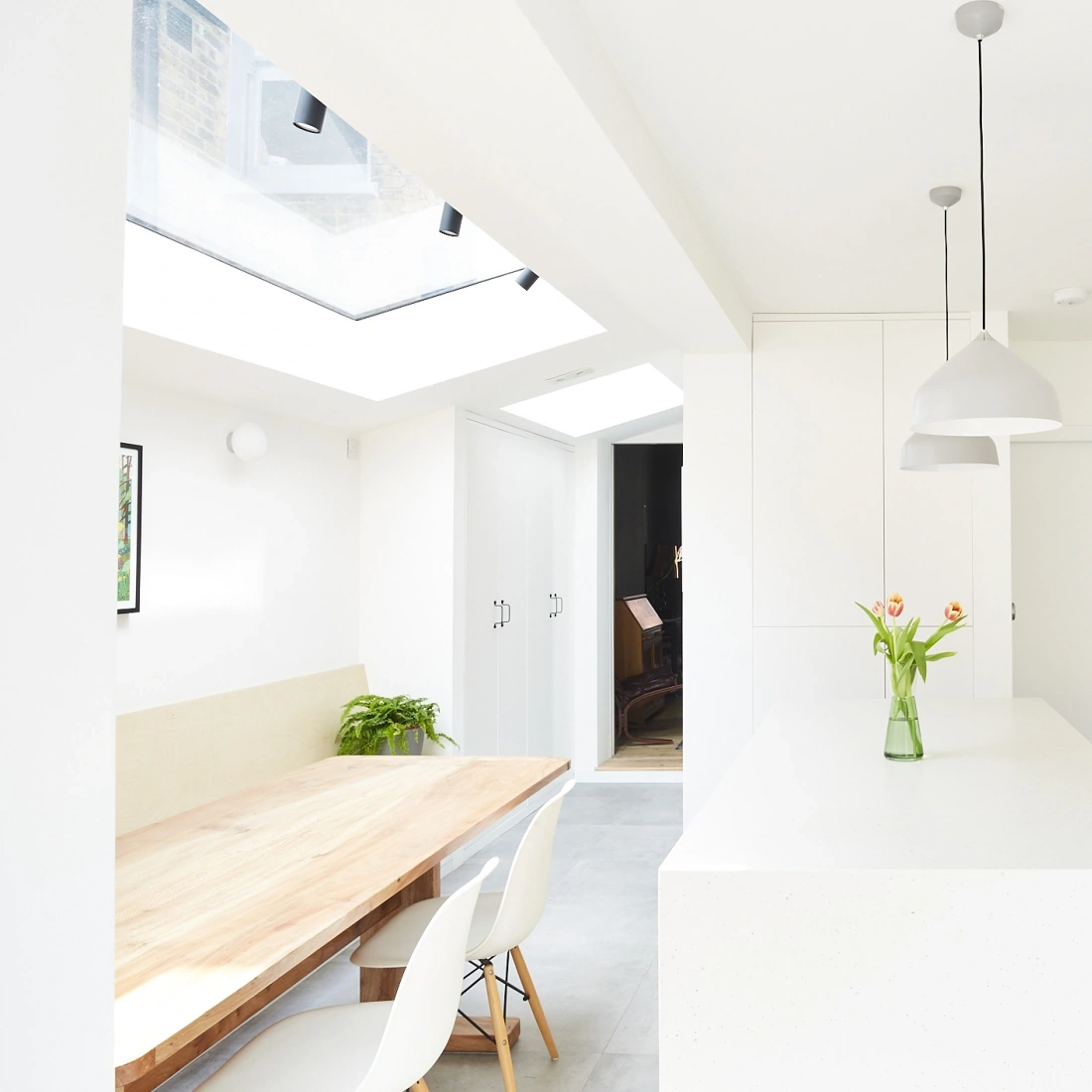 A bright, modern kitchen and dining area featuring a natural wood dining table, white chairs, and a vase of tulips. Large skylights provide ample natural light, highlighting the minimalist decor with white walls and cabinetry.