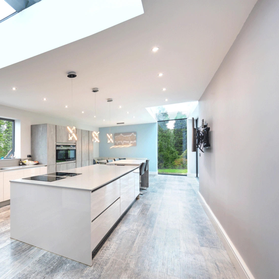 A modern kitchen with a spacious layout, featuring a central island with a sleek countertop. Large windows and a skylight provide ample natural light. The walls are painted in light gray, and the cabinetry has a minimalist design. A wall-mounted TV is visible, along with contemporary appliances.