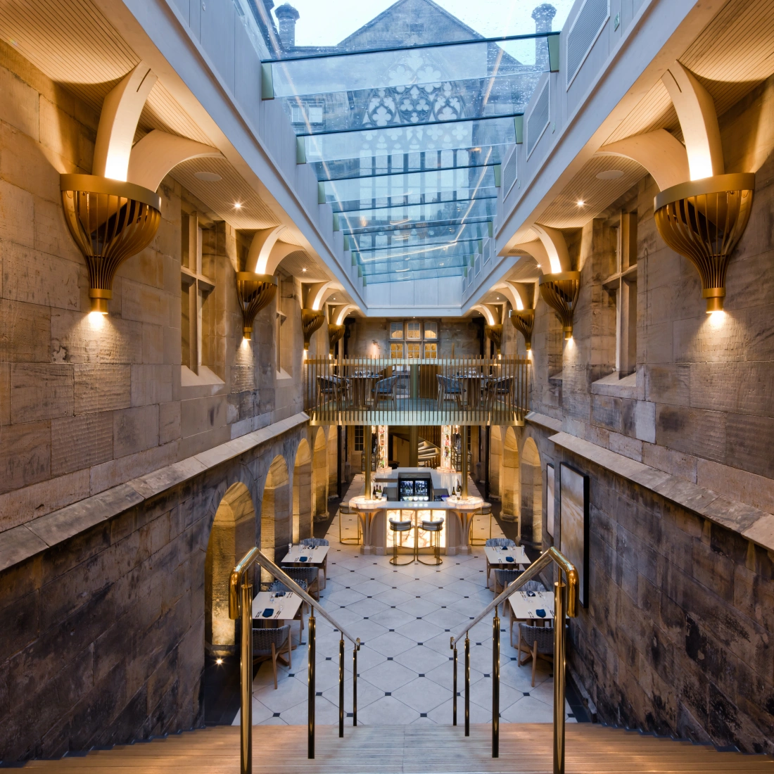 A spacious interior featuring stone walls and elegant architectural details. A grand staircase leads down to a dining area with modern tables and chairs, illuminated by warm lighting. Above, a glass ceiling allows natural light to enter the space.