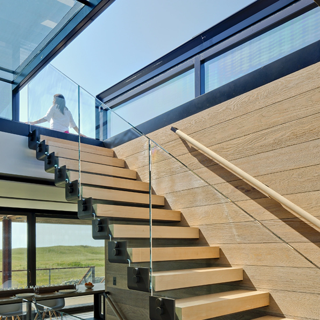 A modern staircase with wooden steps and a glass railing leads up to a bright, open space with large windows. A person is seen ascending the stairs, with sunlight streaming in from above.
