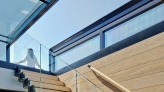 A modern staircase with wooden steps and a glass railing leads up to a bright, open space with large windows. A person is seen ascending the stairs, with sunlight streaming in from above.