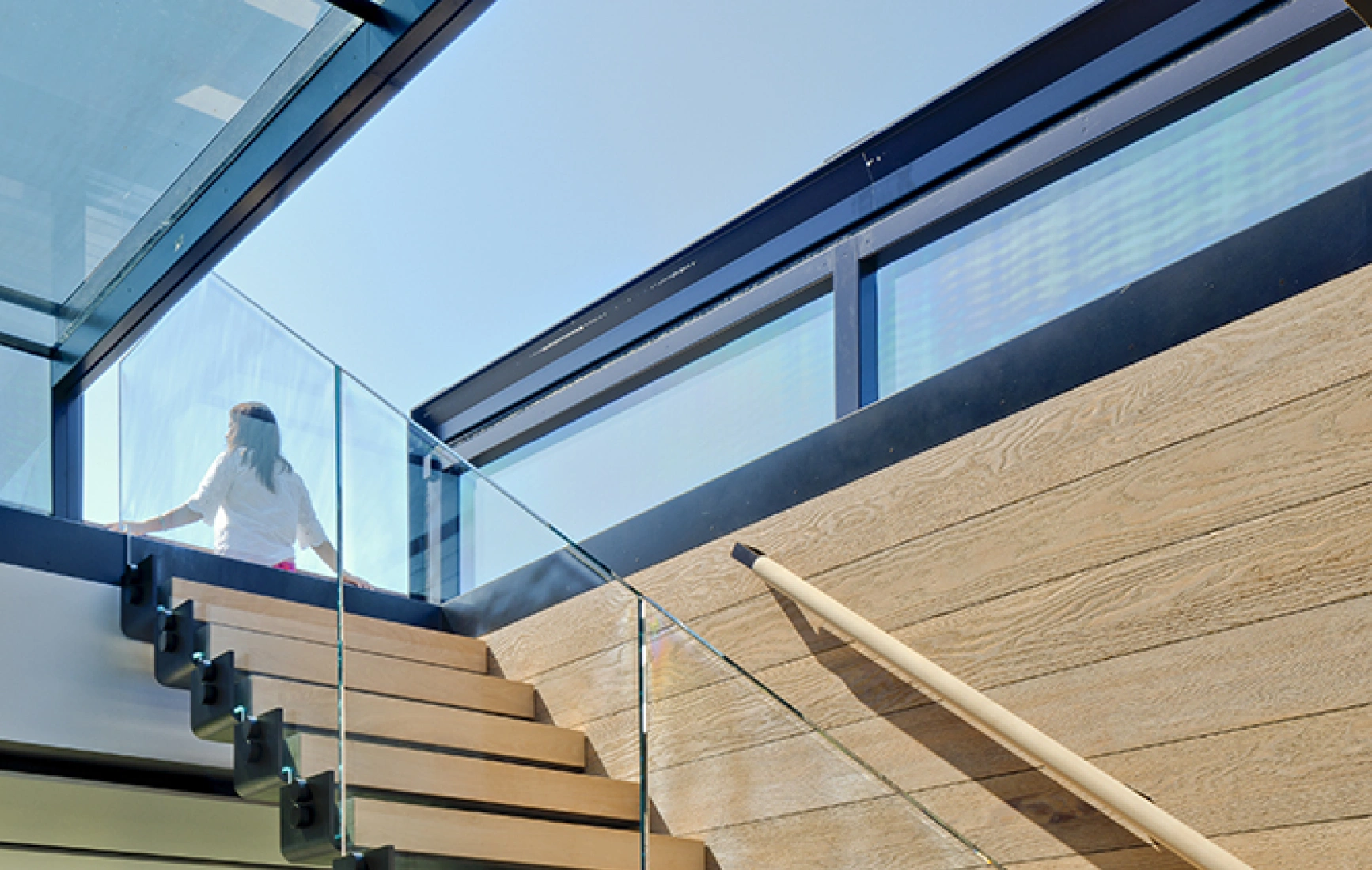 A modern staircase with wooden steps and a glass railing leads up to a bright, open space with large windows. A person is seen ascending the stairs, with sunlight streaming in from above.