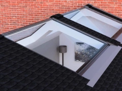 Two roof windows illuminated by natural light, showing an interior space with a gray wall and a lamp, against a backdrop of red brick.