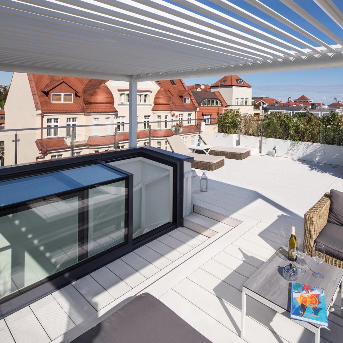 A modern rooftop terrace featuring a wooden deck, lounge furniture, and a view of nearby buildings with red rooftops under a clear blue sky. A table with a bottle of wine and a colorful artwork is displayed.