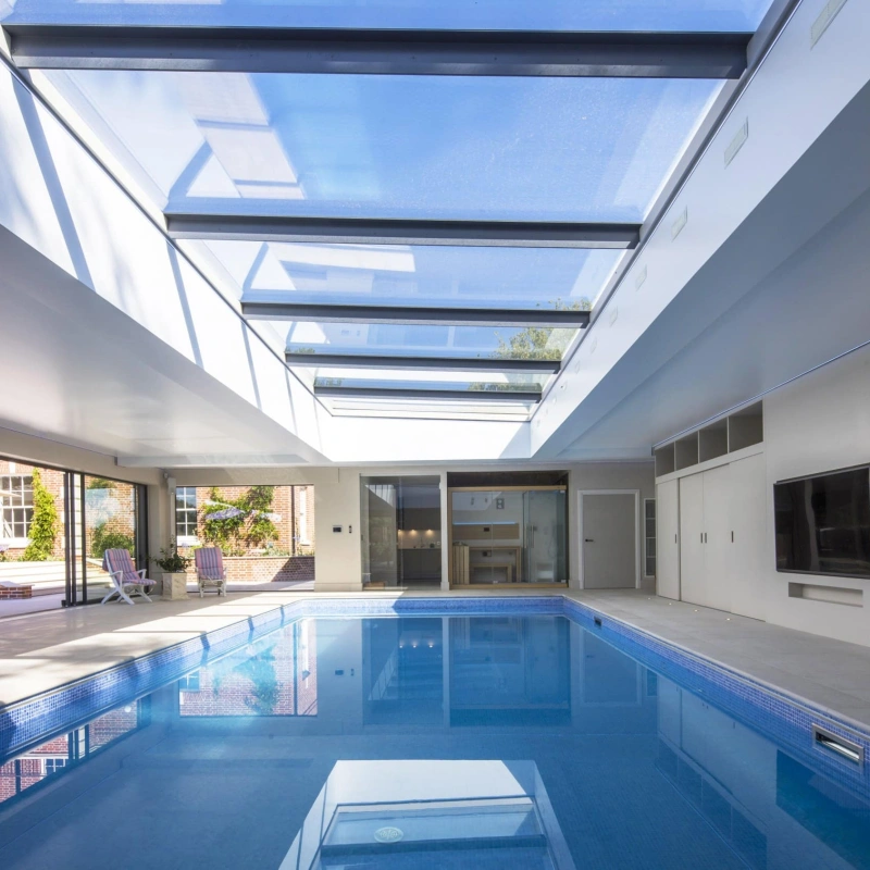 A modern indoor swimming pool features a sleek design with a glass ceiling that allows natural light to flood the space. The pool reflects the surrounding area, which includes sliding glass doors and minimalistic furnishings.
