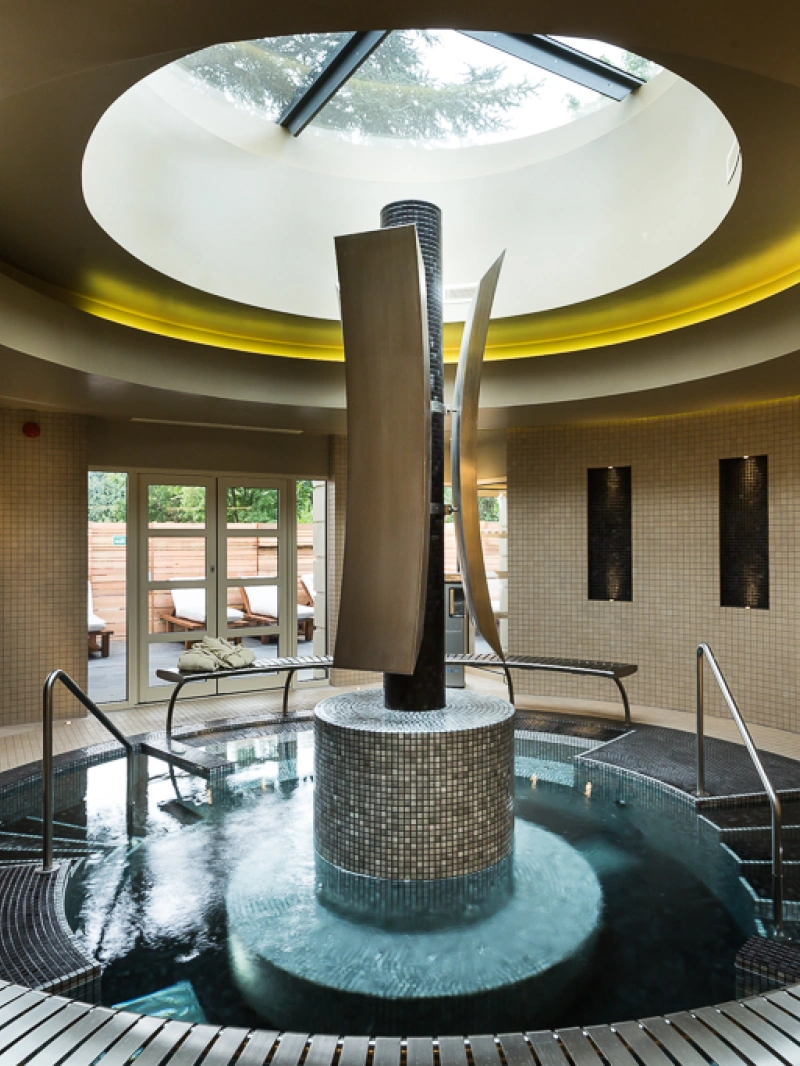 A modern spa interior features a circular skylight and a central water feature with sleek sculptures, surrounded by tiled walls. Comfortable lounge chairs are visible through large glass doors leading outside. Plush towels are neatly arranged on the side.