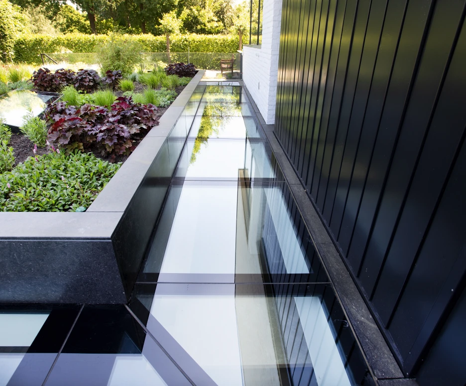 A walkway made of glass panels runs alongside a landscaped area featuring green shrubs and colorful plants. The black exterior of a building is visible in the background.
