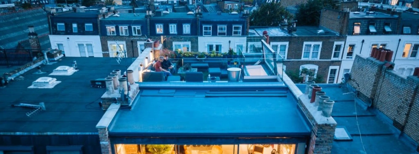 A modern rooftop terrace with a glass-walled lounge area, featuring cozy furniture and decorative plants. Nearby rooftops of city buildings are visible in the background, illuminated by evening lights.