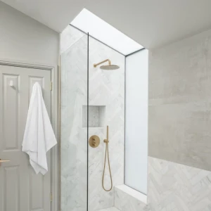 A modern shower with a glass enclosure features a gold showerhead and fixtures. The walls are tiled with white marble in a herringbone pattern, and a window above provides natural light. A folded white towel hangs on the door outside the shower.