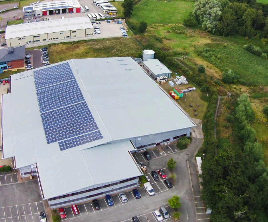 Aerial view of a large commercial building with solar panels on the roof. Surrounding the building are parked cars and a green landscape featuring trees and fields.