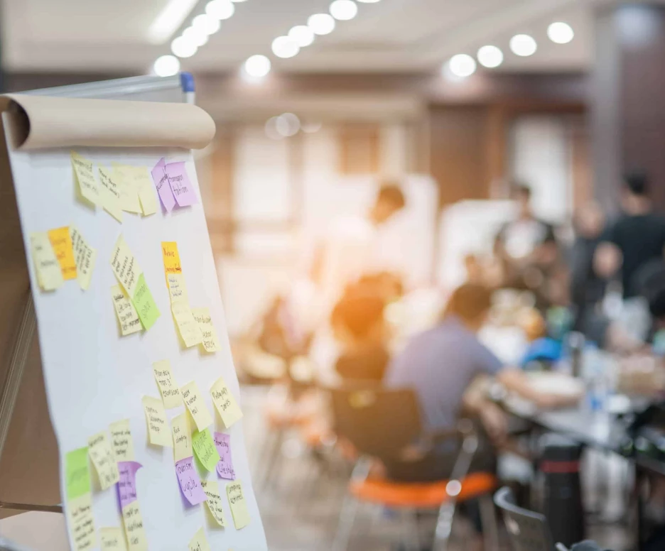 A whiteboard with colorful sticky notes in the foreground, with a blurred group of people engaged in discussion around tables in a well-lit room.