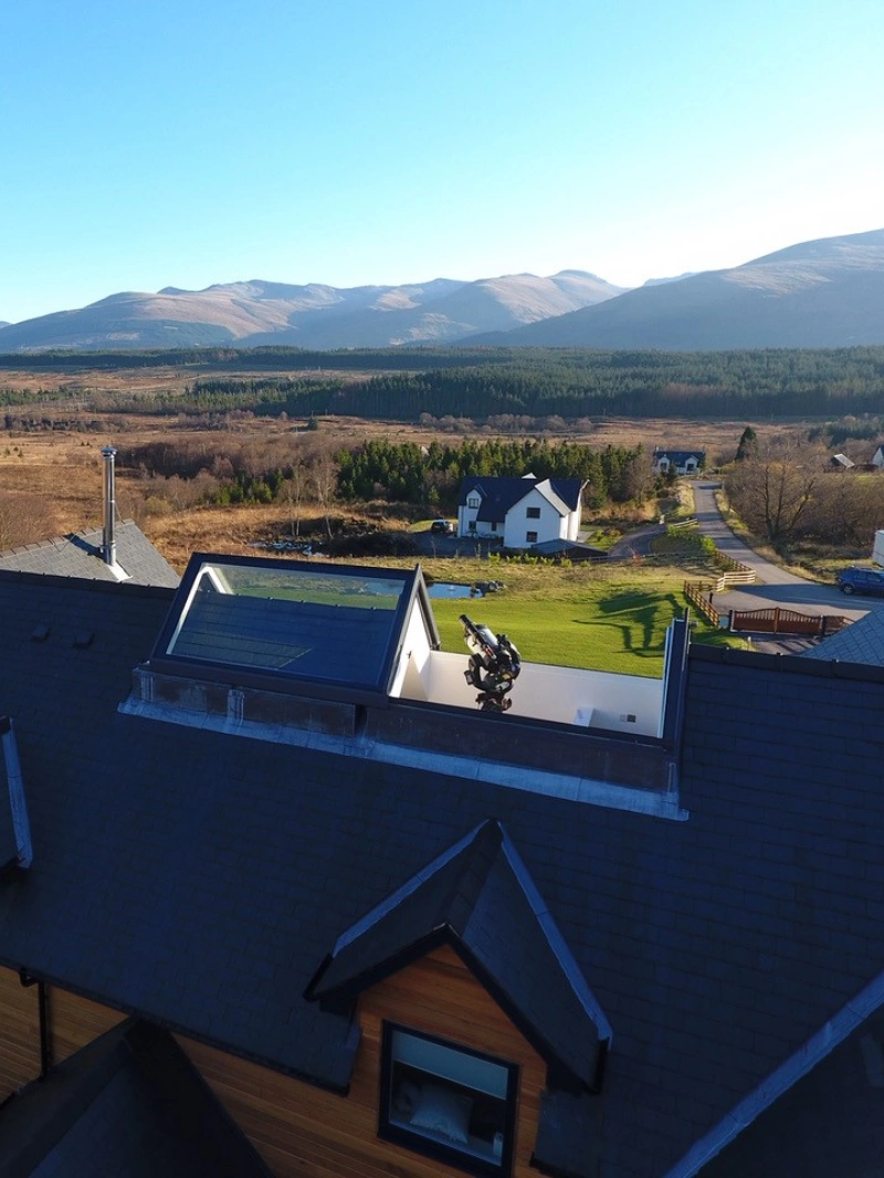Aerial view of a spacious house with a modern design, featuring a sloped roof and large windows. The surrounding landscape includes rolling hills and mountains in the background, with an open field and a few scattered homes nearby. Clear blue sky enhances the serene atmosphere.