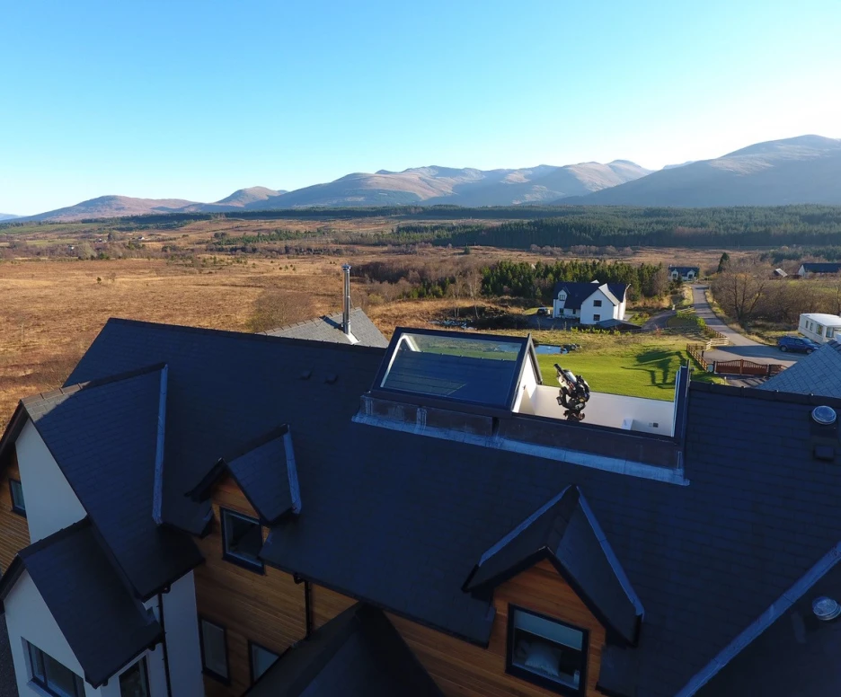 Aerial view of a spacious house with a modern design, featuring a sloped roof and large windows. The surrounding landscape includes rolling hills and mountains in the background, with an open field and a few scattered homes nearby. Clear blue sky enhances the serene atmosphere.