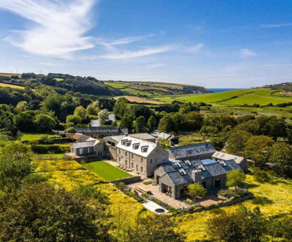 Aerial view of a large stone house surrounded by green fields and trees, with a distant ocean visible under a clear blue sky.
