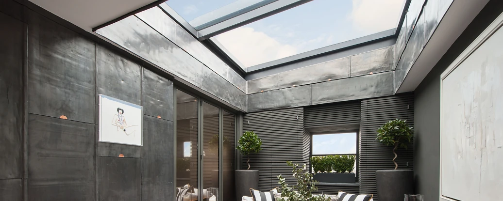 A modern indoor lounge area featuring minimalist furniture, including white sofas with black accent pillows, a round coffee table, and potted plants. Large windows and a skylight provide natural light. The walls are sleek and dark, with artistic decor enhancing the contemporary feel.