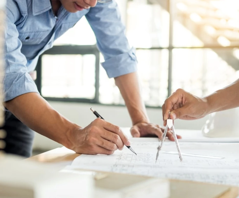 Two people are collaborating on architectural plans, one using a compass and pencil while the other takes notes. A hard hat and construction materials are visible in the background, suggesting a design or construction environment.