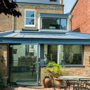 Modernized home extension with a blue metal roof and large glass doors. The structure features a blend of brick and stone, showcasing a spacious outdoor patio with a round table and plants. Sunlight filters in through large windows.