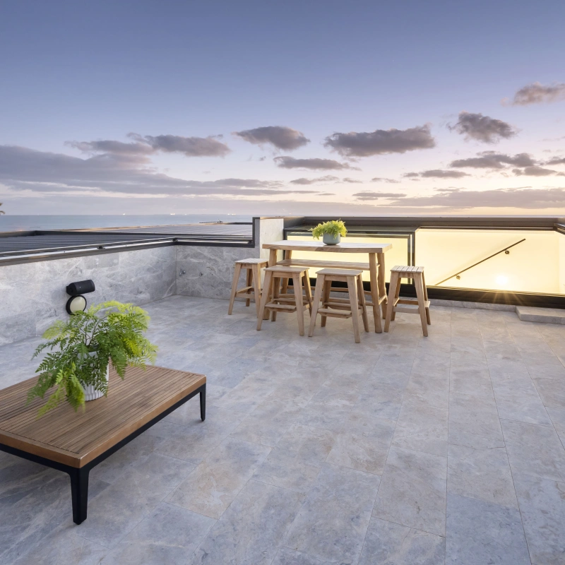 A modern rooftop terrace featuring a wooden table surrounded by bar stools, a low wooden coffee table, and a potted fern. The space offers a view of a serene sunset with clouds in the sky.
